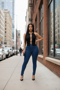 A woman stands on a city sidewalk wearing a black sleeveless top with front cutouts and silver tie details, paired with dark blue skinny jeans and black heels.