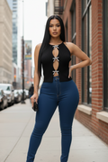 A woman stands on a city sidewalk wearing a black sleeveless cutout top with silver bow details and dark blue skinny jeans, posing with a black clutch.