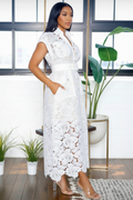 A woman modeling a white lace midi dress with a collar, short sleeves, and pockets, standing in a bright room with plants and large windows.