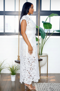 A woman wearing a white lace maxi dress with a high collar and cap sleeves, standing in profile in a bright room with a window, hanging swing, and potted plants.