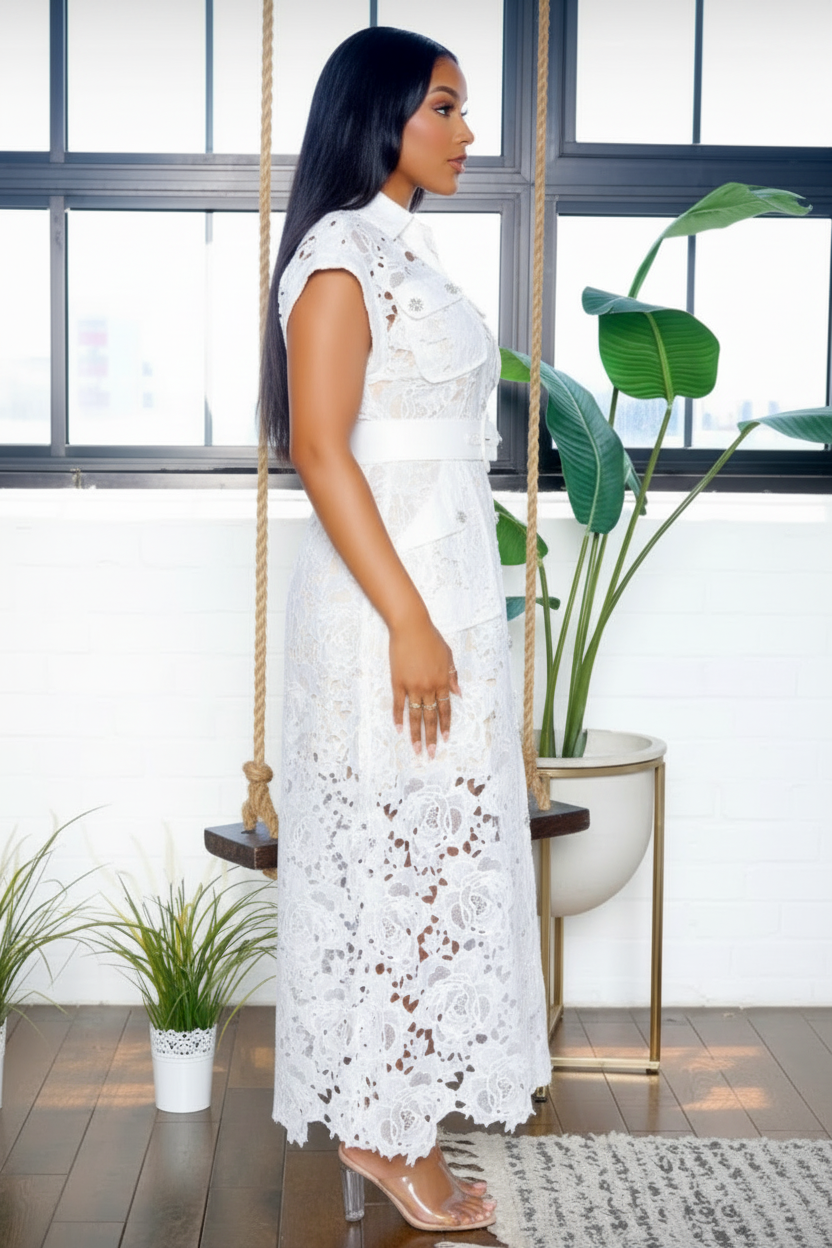 A woman wearing a white lace maxi dress with a high collar and cap sleeves, standing in profile in a bright room with a window, hanging swing, and potted plants.