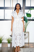 A woman stands indoors wearing a white lace midi dress with short sleeves, a V-neckline, front buttons, and a matching white belt. The setting includes a window, hanging swing, potted plants, and a wooden floor.