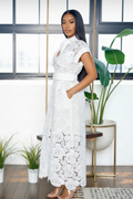 A woman modeling a white lace maxi dress with short sleeves, a high collar, and a matching white belt, standing in a bright room with a hanging swing, potted plants, and a large window.