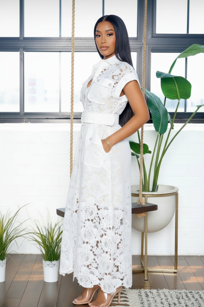 A woman modeling a white lace maxi dress with short sleeves, a high collar, and a matching white belt, standing in a bright room with a hanging swing, potted plants, and a large window.