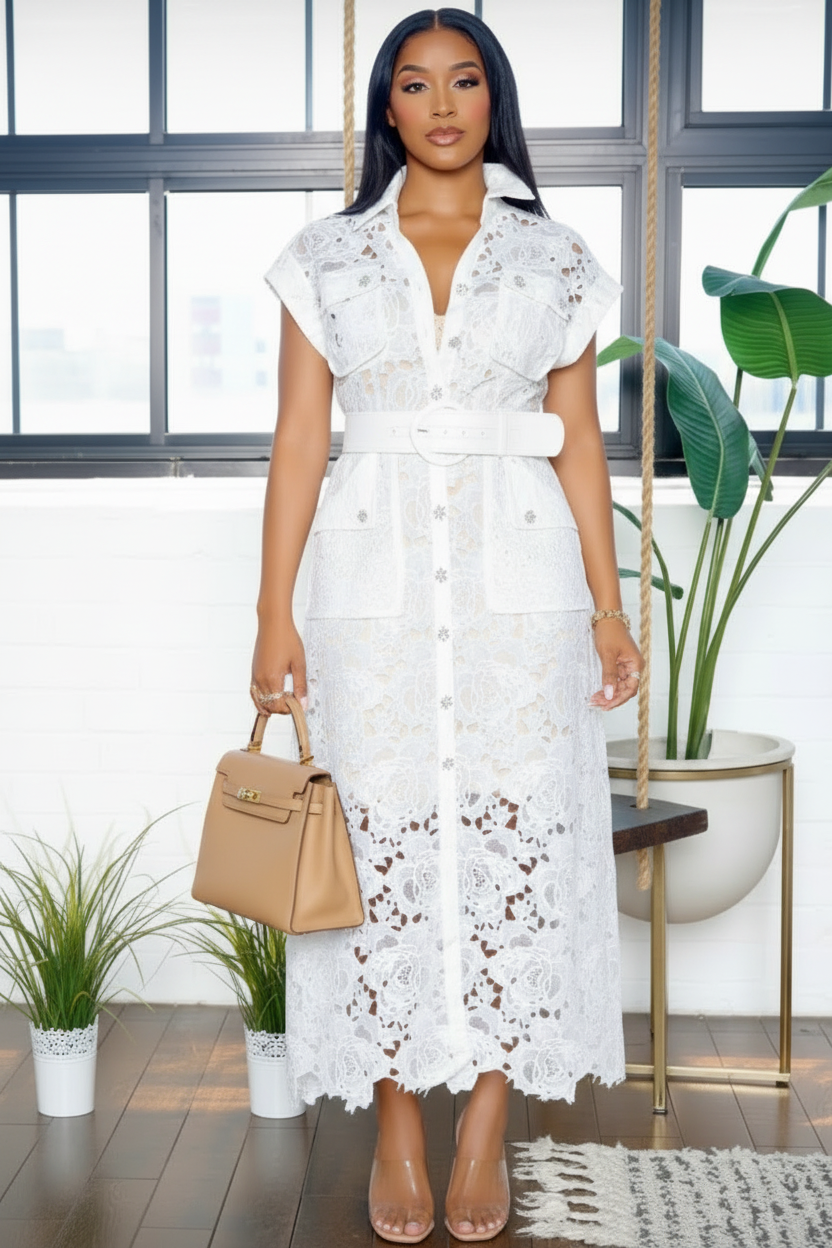 A woman models a white lace midi dress with short sleeves, a button-front closure, and a matching white belt. She is holding a tan structured handbag and wearing clear high-heeled sandals in a bright room with large windows and potted plants.