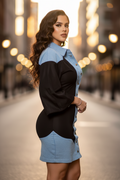 A woman posing in a color-block denim shirt dress with a light blue collar, sleeves, and skirt panels, and a black body, set against a blurred city street at dusk.