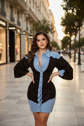 A woman stands on a city sidewalk wearing a color-block mini dress with a light blue denim-like collar, sleeves, and hem over a black body. The dress has a front button placket and is photographed in an outdoor urban setting with buildings and trees in the background.