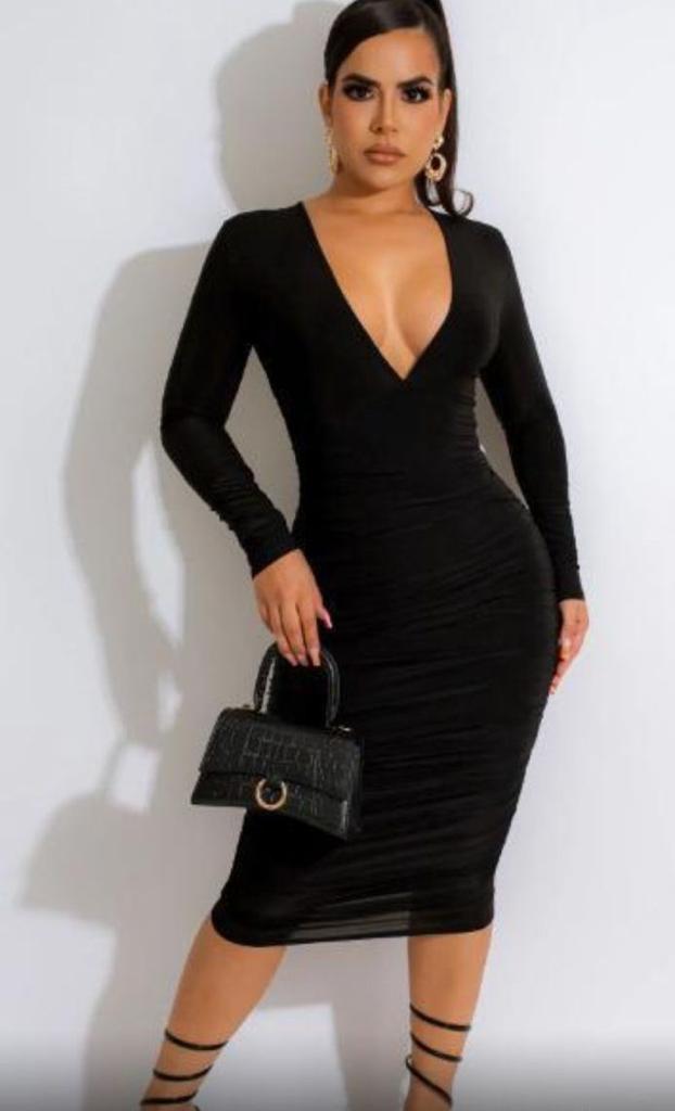 Woman wearing a black dress with long sleeves and a deep neckline, holding a black handbag against a white background.