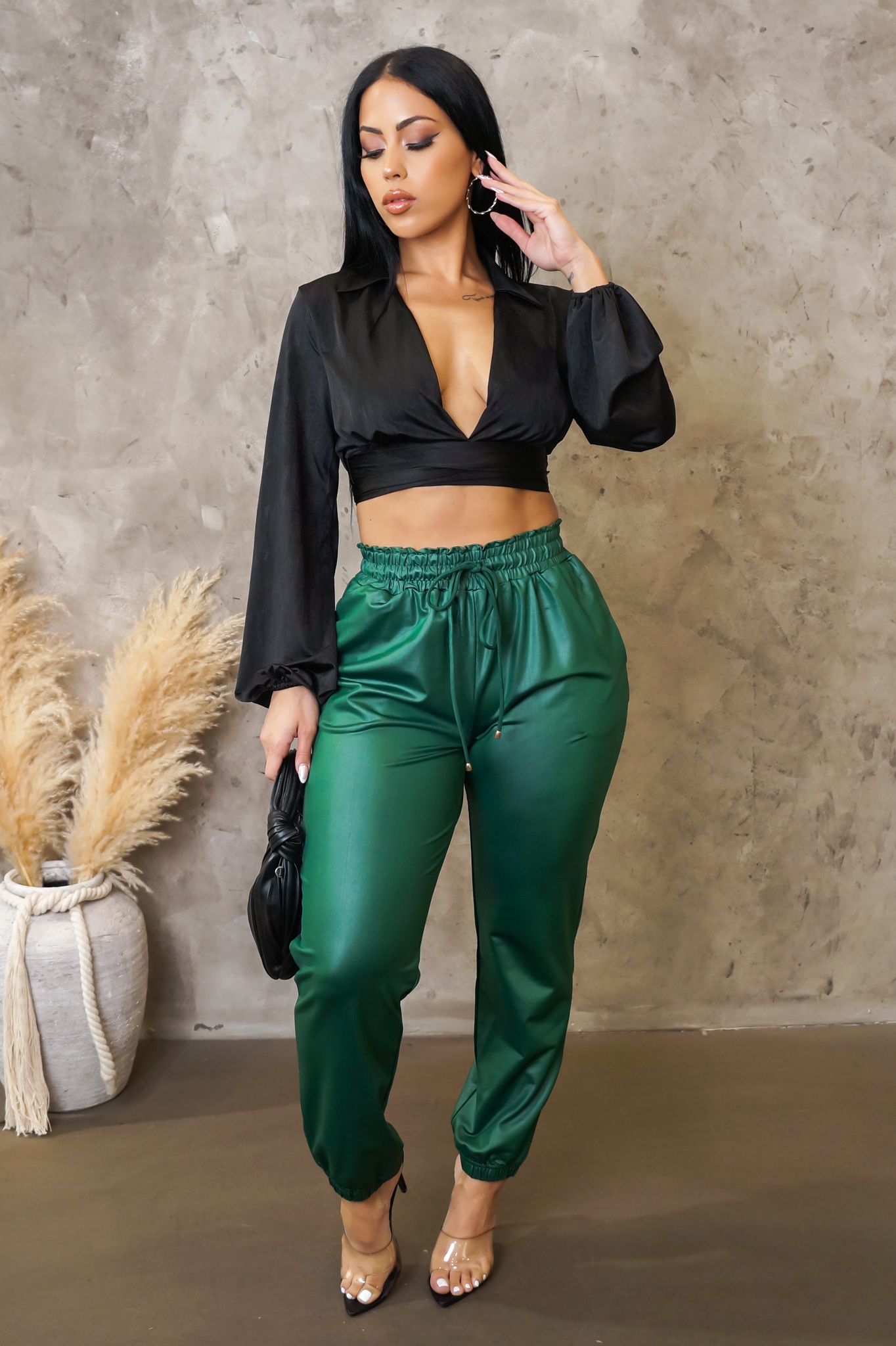 Woman wearing a black top and green pants against a concrete wall.